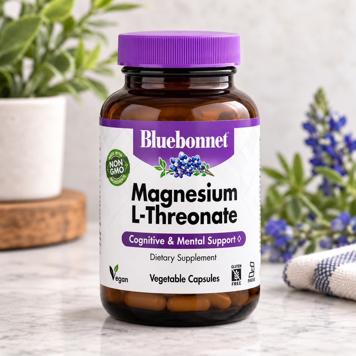 A bottle of Bluebonnet Magnesium L-Threonate dietary supplement capsules, set on a marble surface with blurred potted plants and flowers in the background.