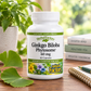 A bottle of HerbalFactors Ginkgo Biloba Phytosome dietary supplement capsules, placed on a wooden table with a potted plant, books, and a notebook in a bright, natural setting.