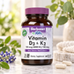A bottle of Bluebonnet Vitamin D3 & K2 dietary supplement, 60 veggie capsules, set on a marble surface with blurred green leaves and purple flowers in the background.